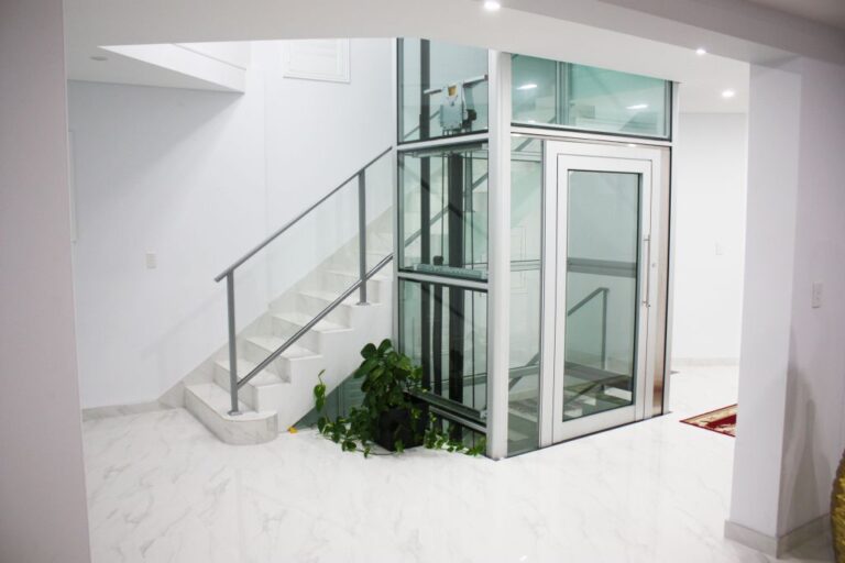 Glass shaft home lift beside a white marble staircase with a glass balustrade in Los Angeles