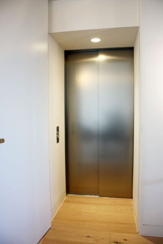 Narrow lift door with a brushed steel finish at the end of a timber-floored passage in a Fitzroy home