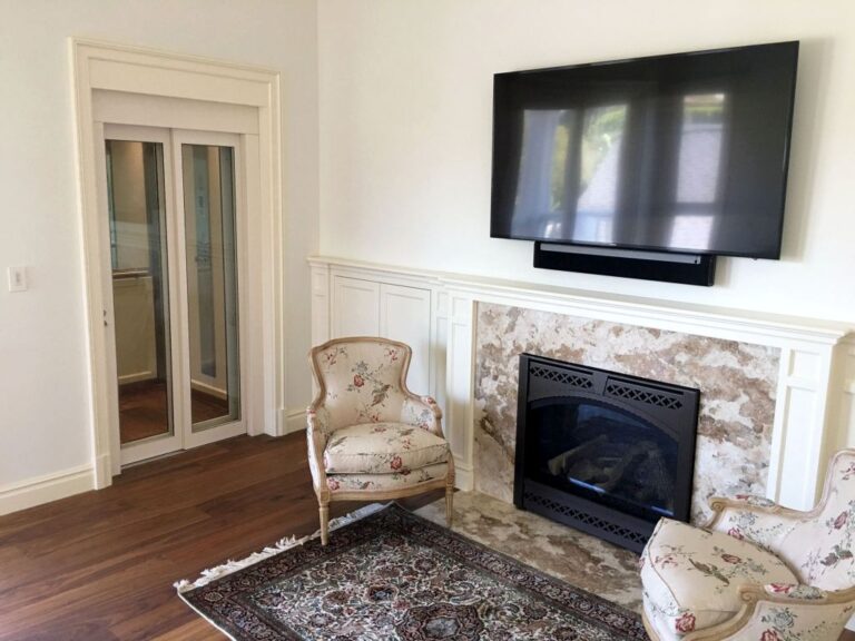Glass panelled lift door in a white frame beside a fireplace with floral armchairs in Hollywood