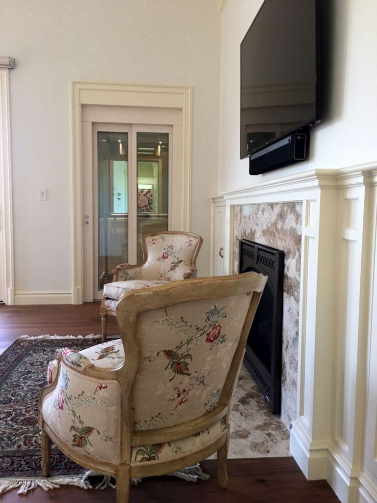 Glass-panelled lift door in a cream architrave behind floral armchairs and a marble fireplace