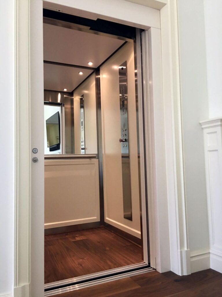 Open E2 lift cabin with cream panels, brushed stainless steel trim, a mirror and a dark hardwood floor
