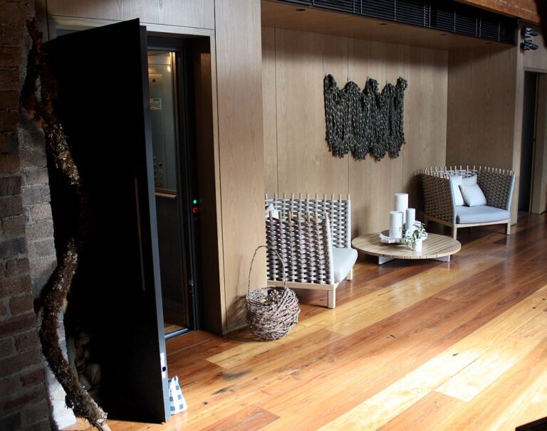 Open black lift door beside a timber wall and lounge chairs on a timber floor