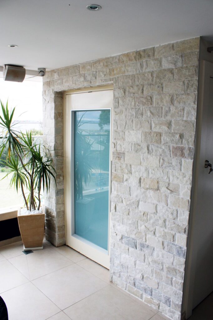 Milk white glass lift door in a stone-clad wall beside a potted palm and a bay view