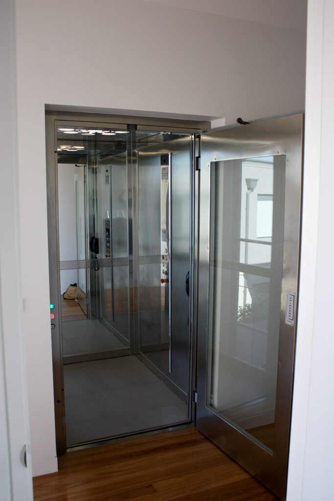 Open stainless steel and glass lift door showing the mirrored, brushed stainless steel cabin and lit ceiling diffuser in a Lane Cove home