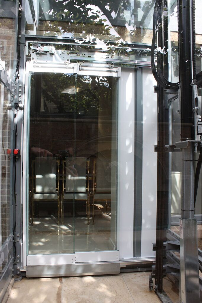 Looking into the glass shaft lift cabin with a mirrored back wall, framed by the shaft steelwork