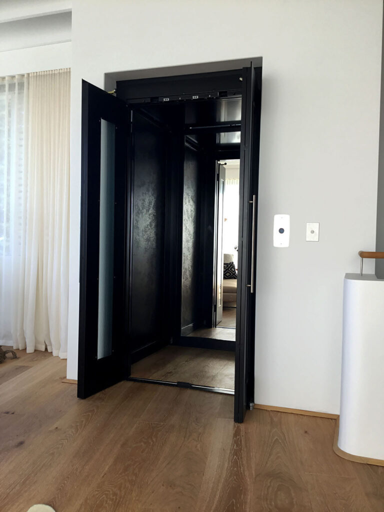 Black lift door open to a mirrored Arpa Black cabin beside a white wall and timber floor