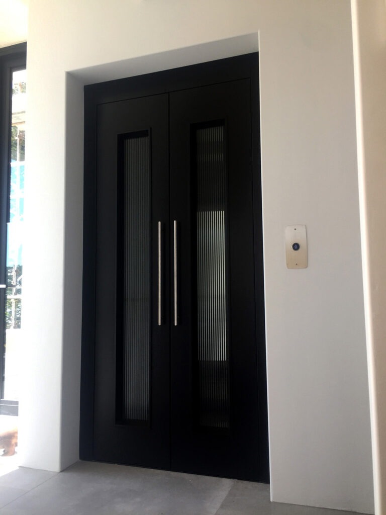 Closed black double lift doors with fluted glass panels and long steel handles set in a white wall