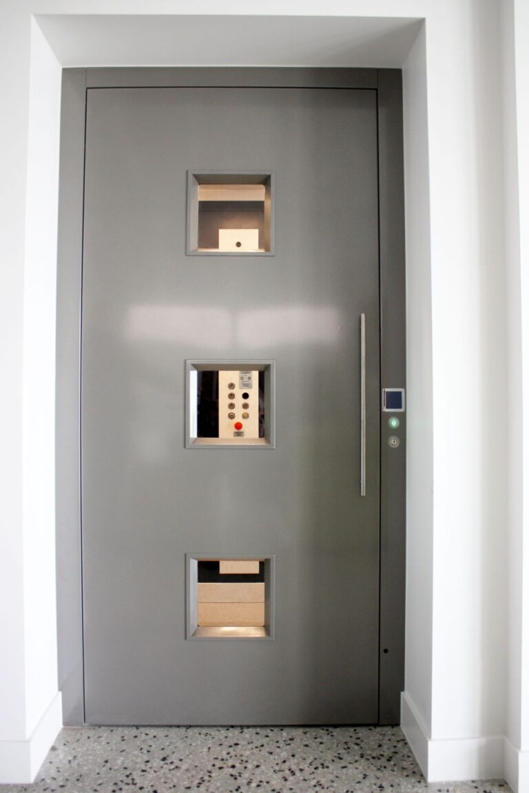 Closed grey lift door with three small square windows and a long steel handle