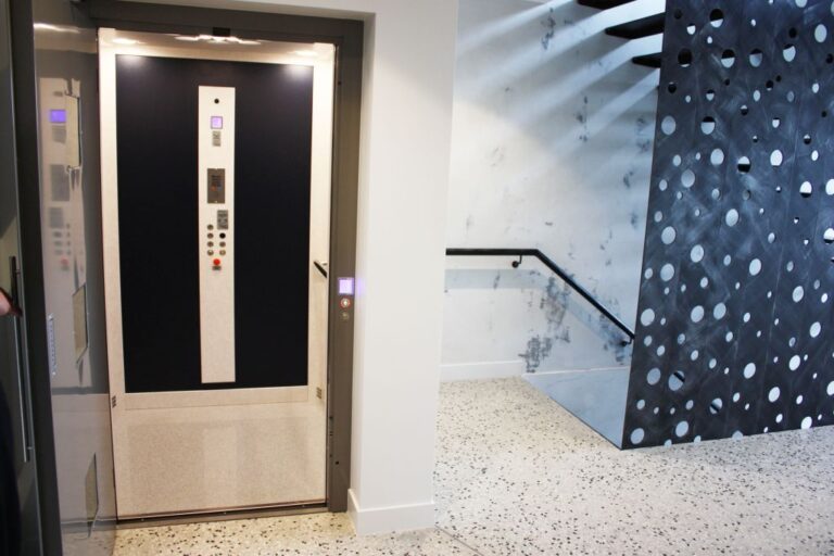 Open lift door showing a black panel with a button strip on a speckled terrazzo floor