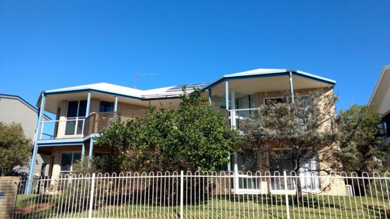 Two storey brick home with balconies and a white fence under a blue sky on the Sunshine Coast