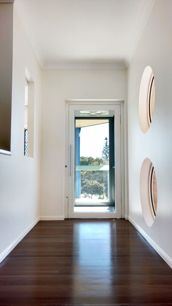 Hallway with a timber floor, two round windows and the glass lift door at the end