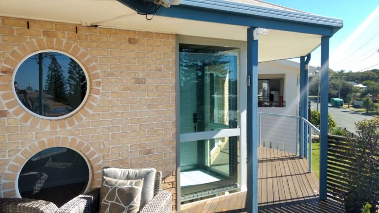 Glass lift with a blue frame beside round windows on a brick wall and deck seating