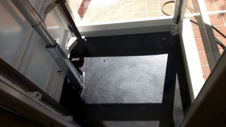 Looking down into the glass lift shaft pit from the top, with the rails and drive beside it