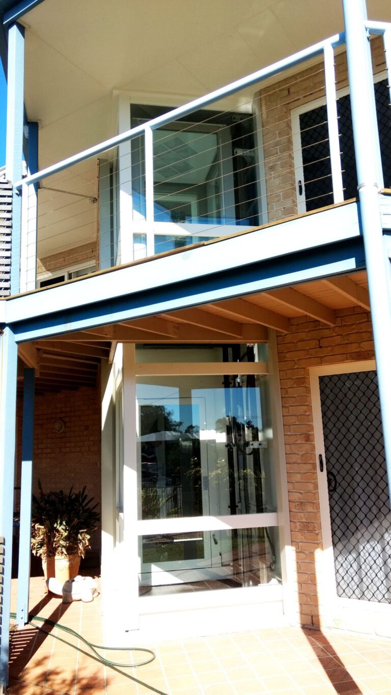 Two-level glass lift tower beside a brick wall under a balcony with a wire balustrade on the Sunshine Coast