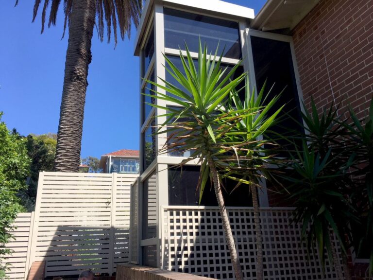 Glass lift shaft rising beside a brick wall and palm trees in Mosman
