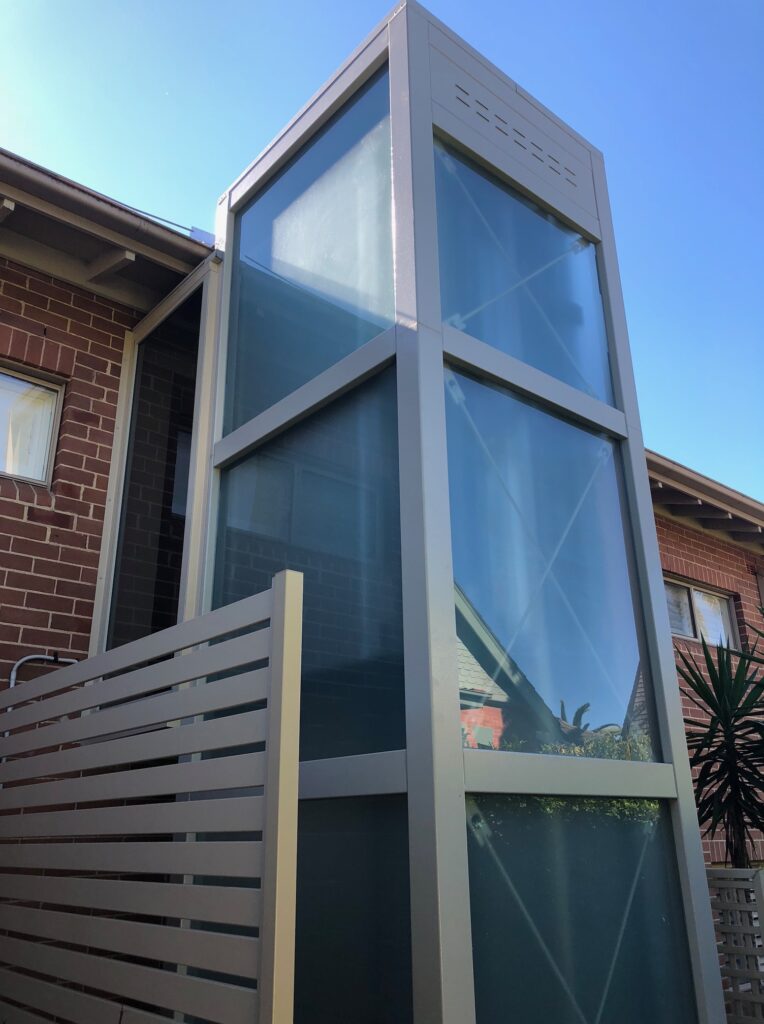 Glass lift shaft with a slatted privacy screen against a brick home