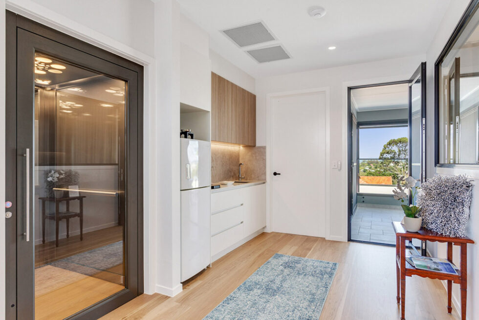 Dark-framed glass lift door beside a kitchenette with timber cabinets and a balcony beyond, in a two-storey Queensland home