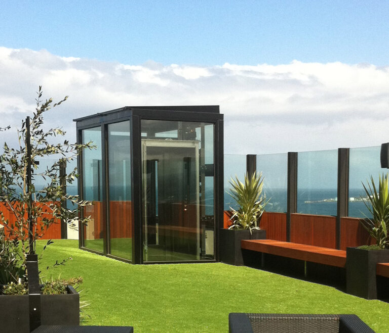 Glass lift enclosure on a rooftop lawn with ocean views behind a glass balustrade