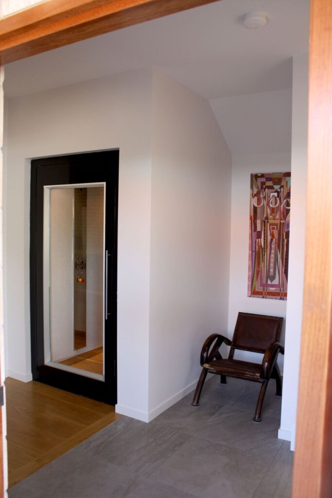 Black framed glass lift door on a white landing with a timber floor and a chair beside it