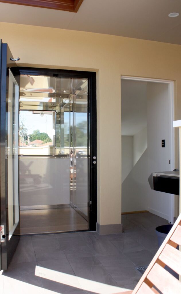 Black framed lift door open at the rooftop level, showing the cabin and neighbourhood rooftops through the glass