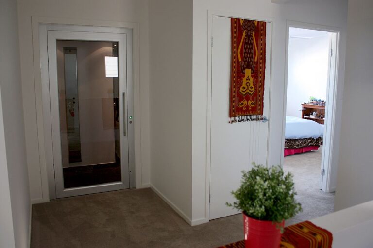 Glass-panelled lift door on the carpeted upper landing beside a bedroom doorway