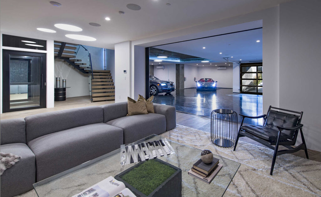 Living room with a grey sofa and an open garage beyond, with the black-framed glass E1 lift door at left beside floating stairs