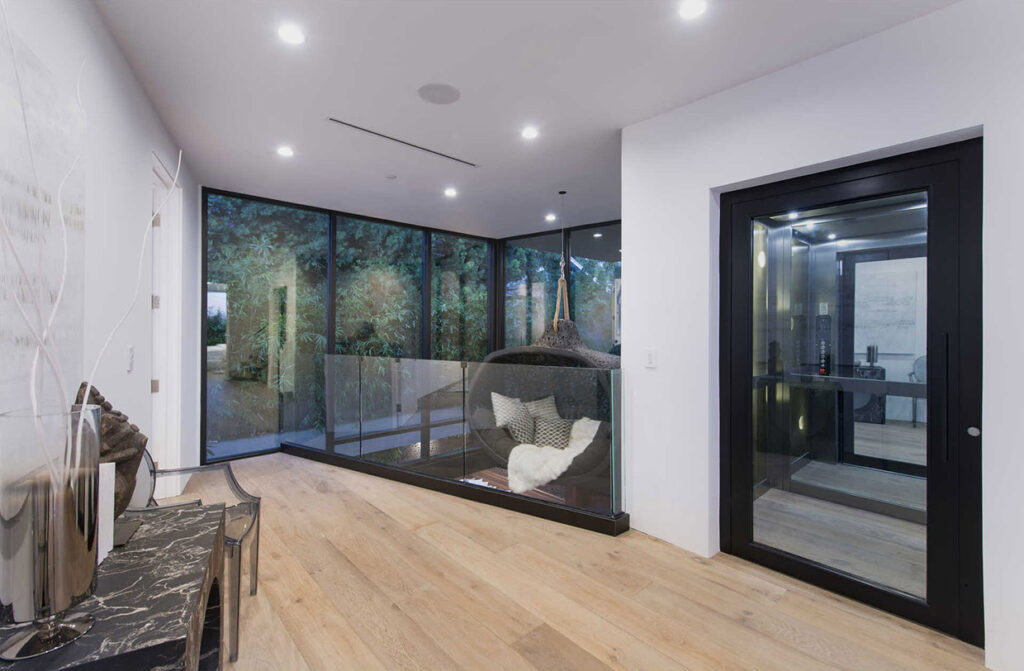 Upper landing with a black-framed glass E1 lift door and timber floor beside floor-to-ceiling windows