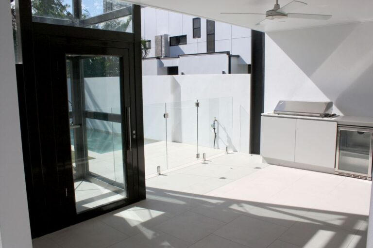 Black framed glass lift door opening to a patio with a barbecue and pool fence