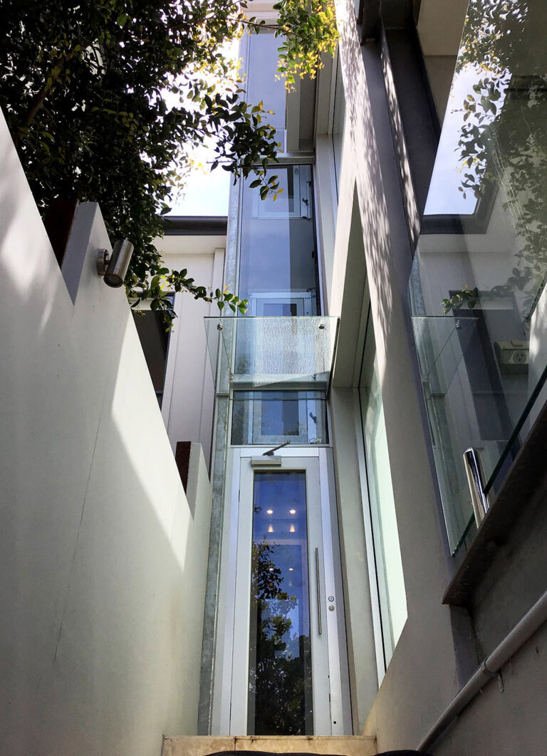 Narrow glass lift shaft between white walls with greenery above in Fairfield
