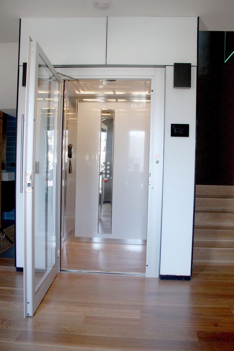 Open glass lift door in a white shaft on a timber floor beside the stairs