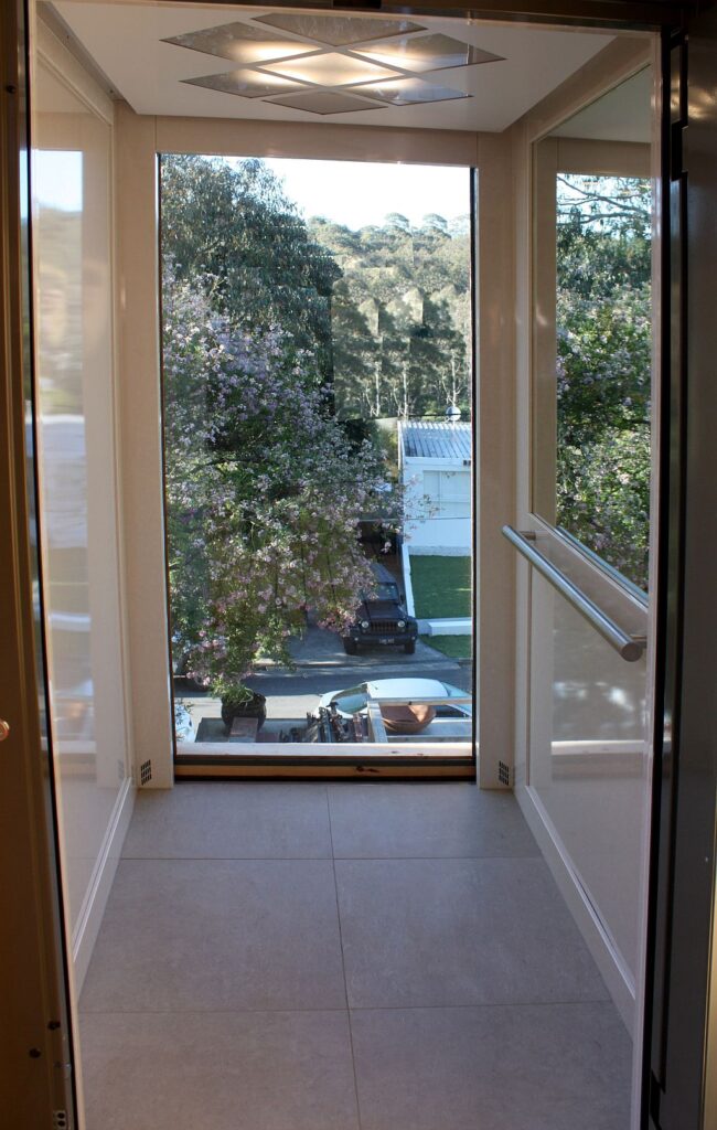 Looking through the lift car to the garden beyond the glass door, with a stainless steel grab rail