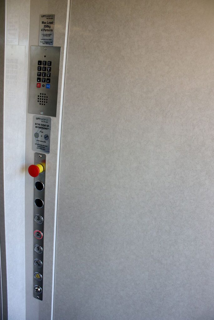 Lift car operating panel with buttons, an emergency stop and a telephone beside a pale skinplate wall