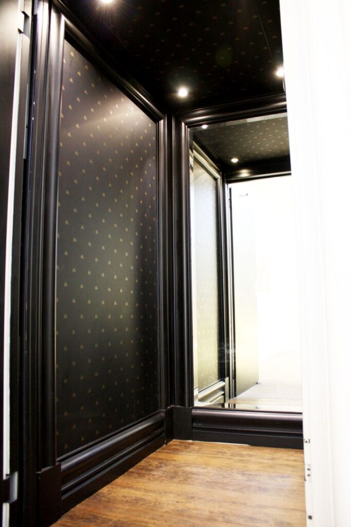 Inside the E1 lift cabin: black-painted timber mouldings, patterned wallpaper, a full-height mirror and a hardwood floor