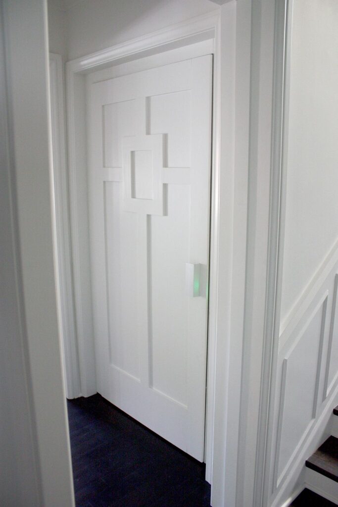 White clad lift landing door with a square panel pattern and a bespoke handle, beside the staircase