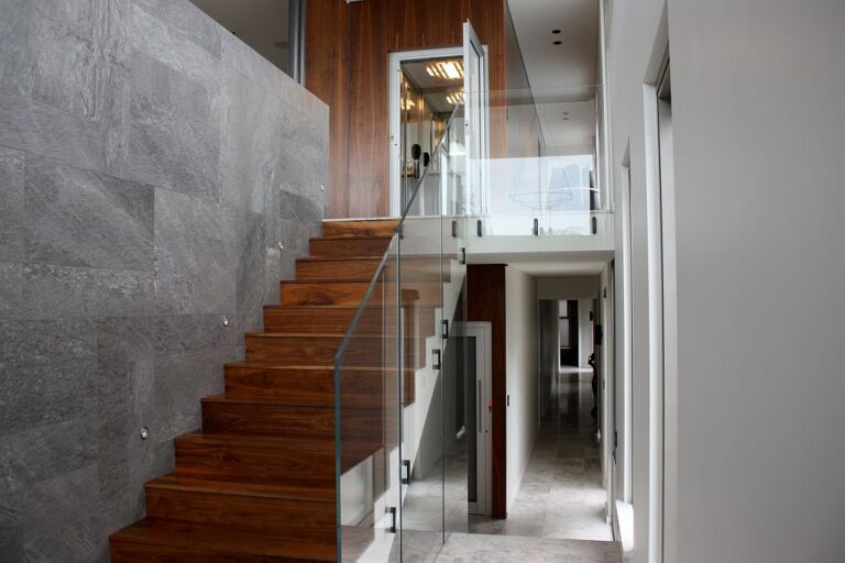 Timber staircase with glass balustrade beside a grey stone wall and a lift door