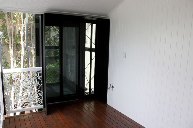Black-framed glass lift doors on the upper verandah landing of a Queenslander, beside the white fretwork balustrade