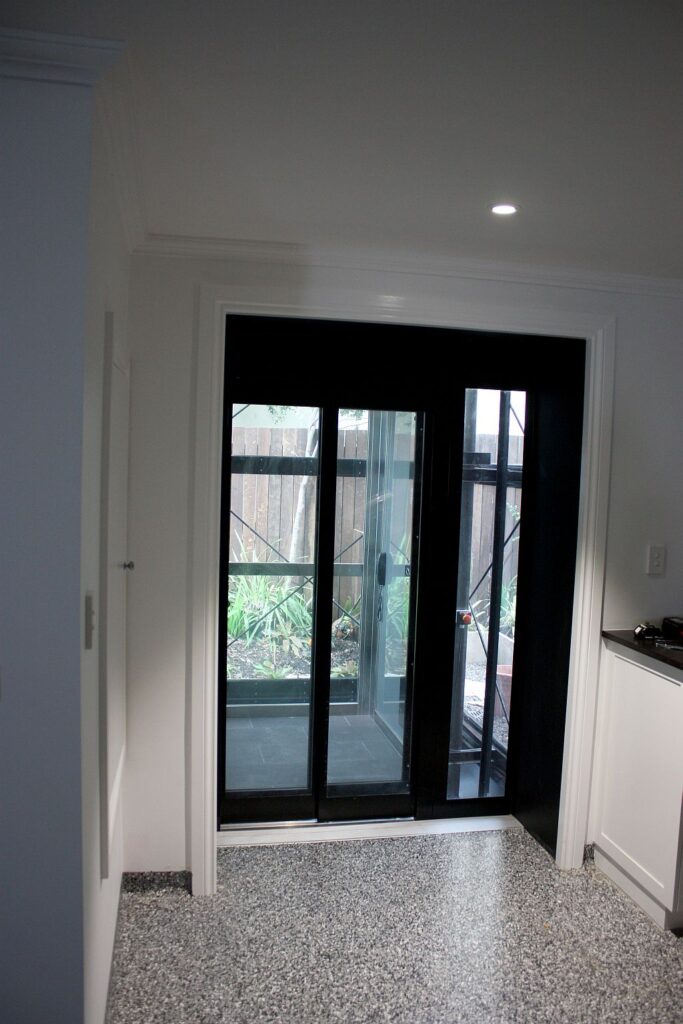 Glass and black lift doors opening into a hallway, with the garden visible through the glass cabin