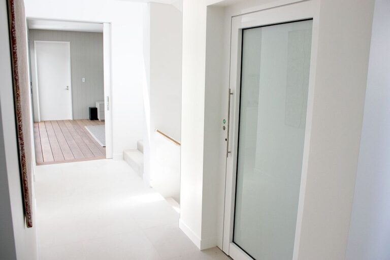 Lift door with a frosted glass panel in a white hallway beside a timber deck