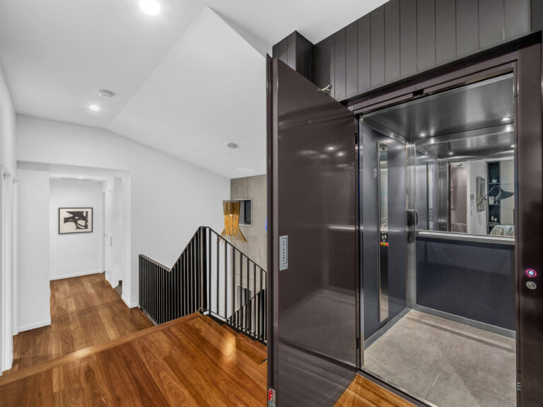 Open lift door in a dark panelled wall beside a timber stair landing in Coorparoo