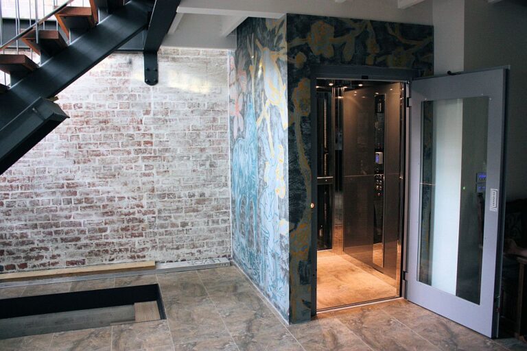 Open lift door beside an exposed brick wall and patterned wallpaper in a converted factory