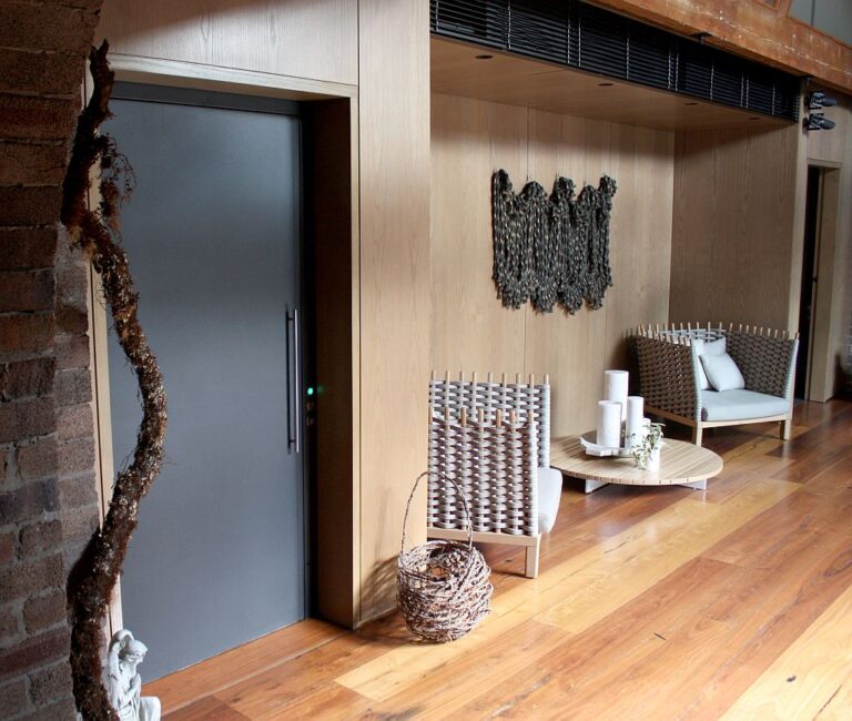 Grey lift door beside a timber panelled wall with a woven art piece and lounge chairs
