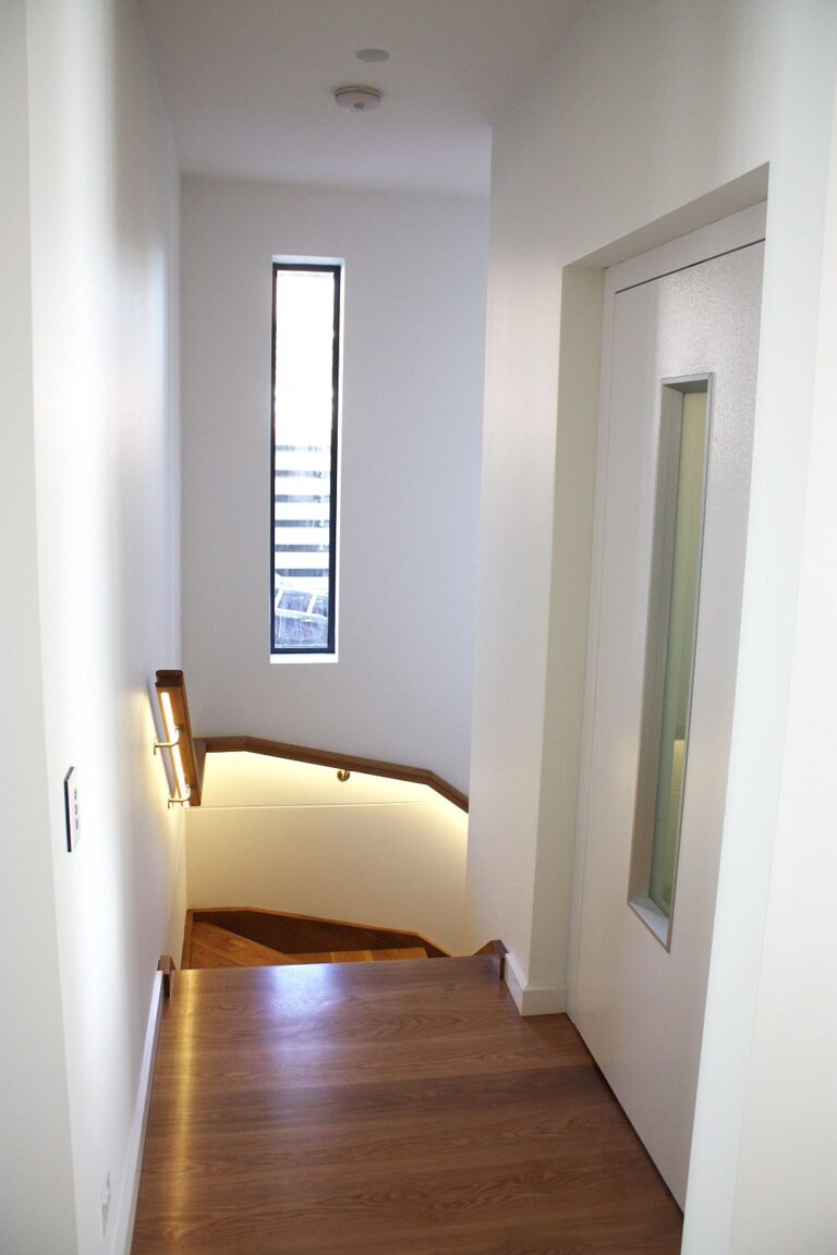 Hallway with a white lift door beside a timber staircase with strip lighting in a Sydney home