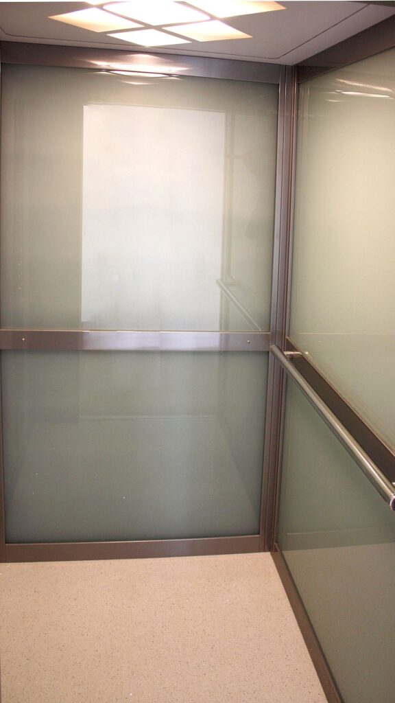 Inside the ElfoLift cabin, with milk glass walls, a stainless handrail, a stone floor and a square ceiling light