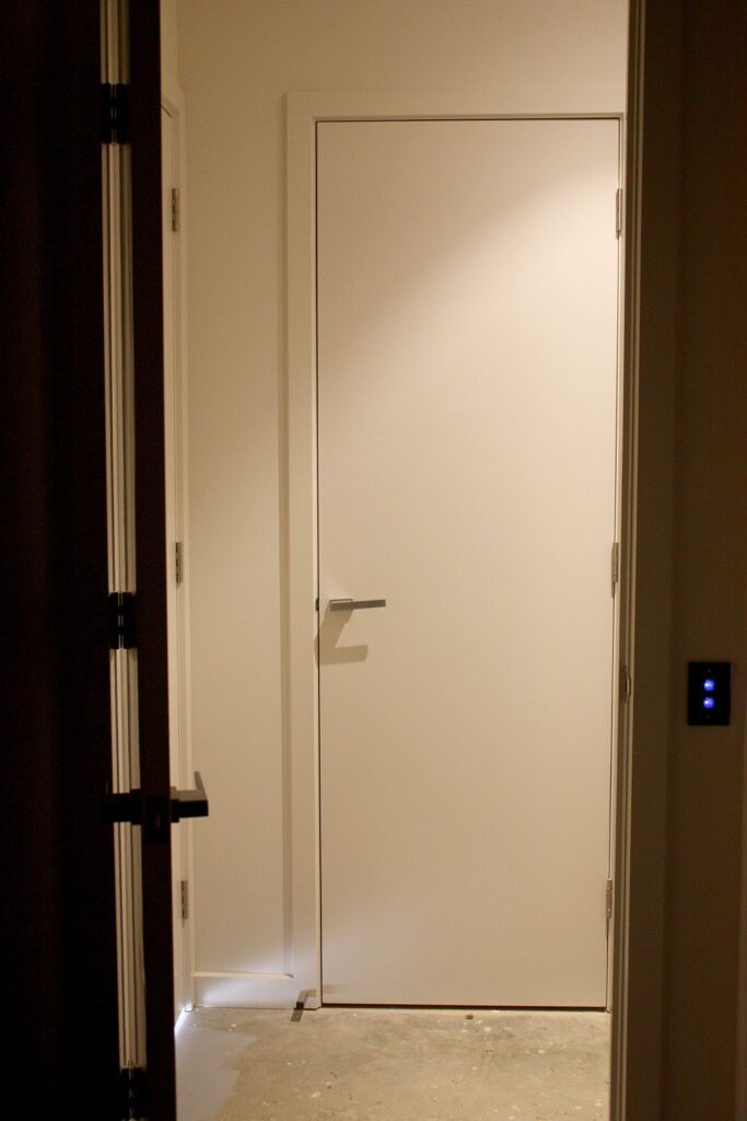 Flush white lift landing door in a dark frame beside the call buttons, with the floor still bare concrete