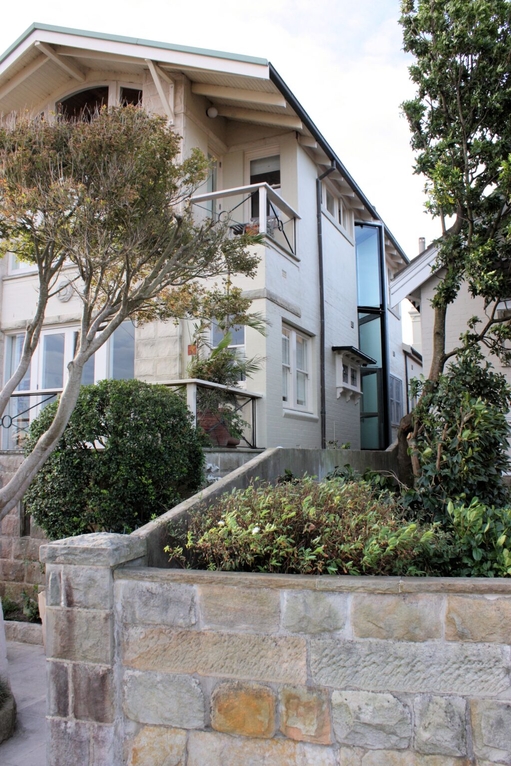 Glass lift shaft rising up the side of a Mosman house behind a sandstone garden wall