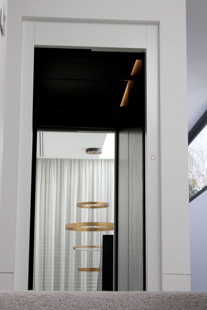 White-framed lift landing door with the black cabin and a gold ring chandelier reflected inside
