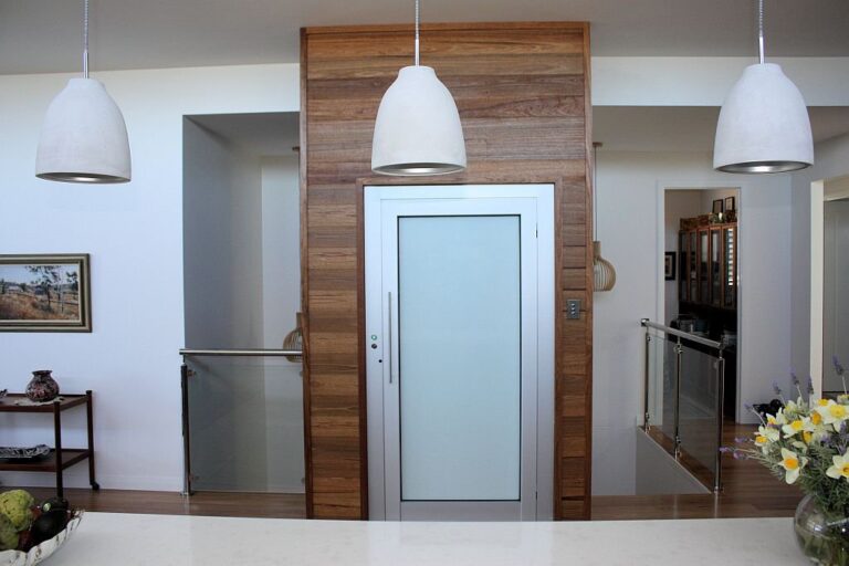 Frosted glass lift door set in timber cladding under three pendant lights, with glass balustrade beside it