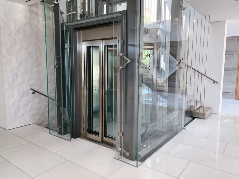 Front view of a glass walled lift with a stainless steel door and textured white wall