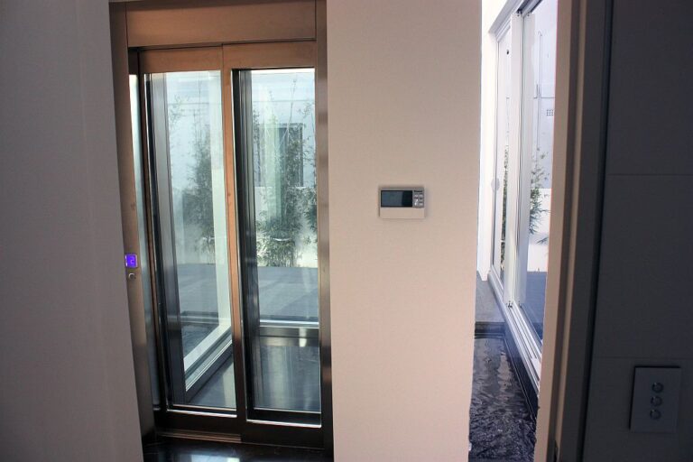 Stainless steel and glass landing doors beside a wall control, with the courtyard pond visible beyond