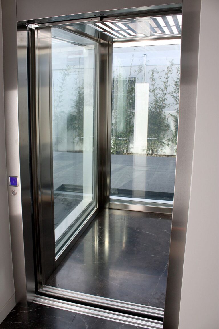 Open E2 lift car with glass walls, brushed stainless steel frame and brown marble floor, courtyard bamboo visible through the glass